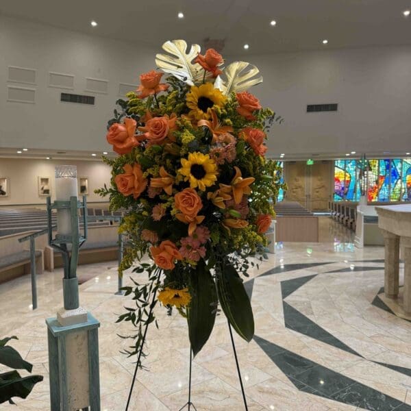 Orange and gold funeral standing spray with roses, sunflowers, lilies and gold tropical leaves in Naples Florida.